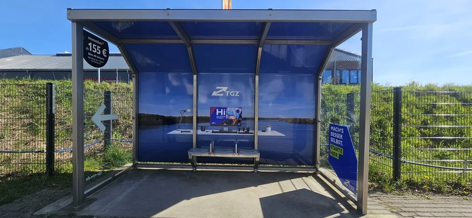 Gläserner Warteunterstand mit blauer TGZ-Werbegestaltung und Sitzbank